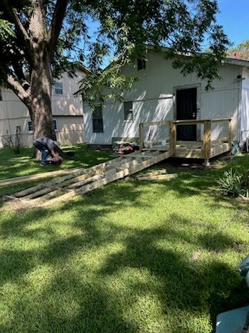 A volunteer bent over a saw in a sunlit back yard while the ramp behind him gets its decking and handrail posts.