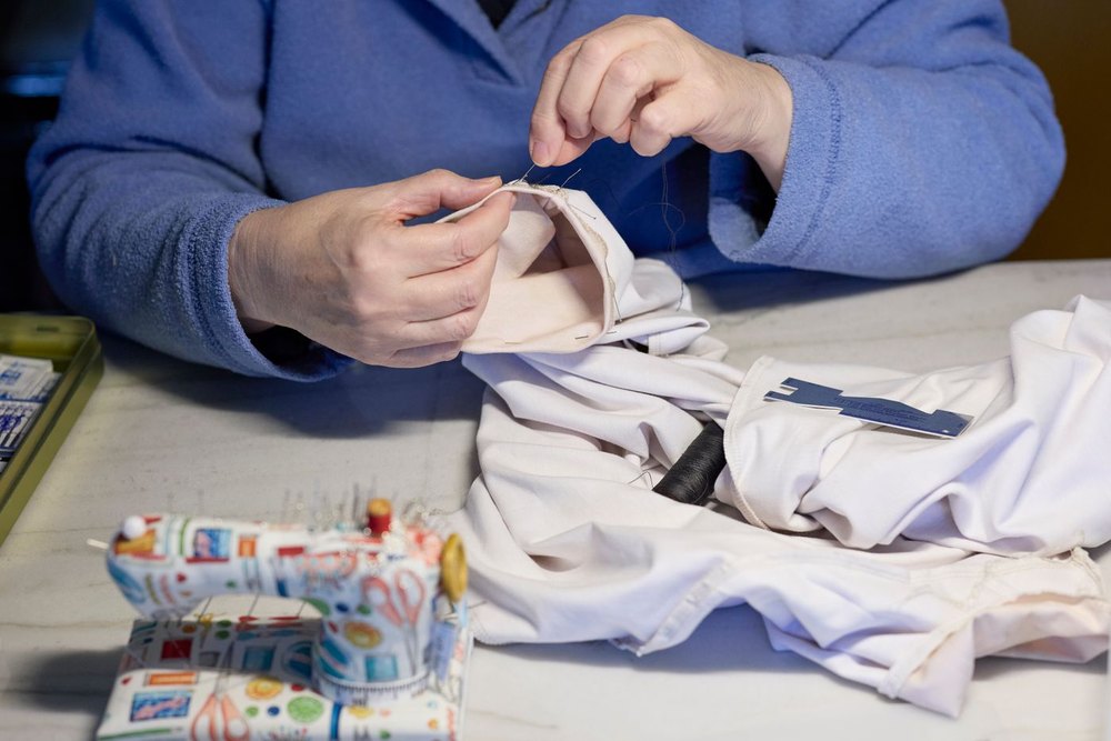 A woman at a work table hand-sewing a white garment, with a sewing box open beside her.